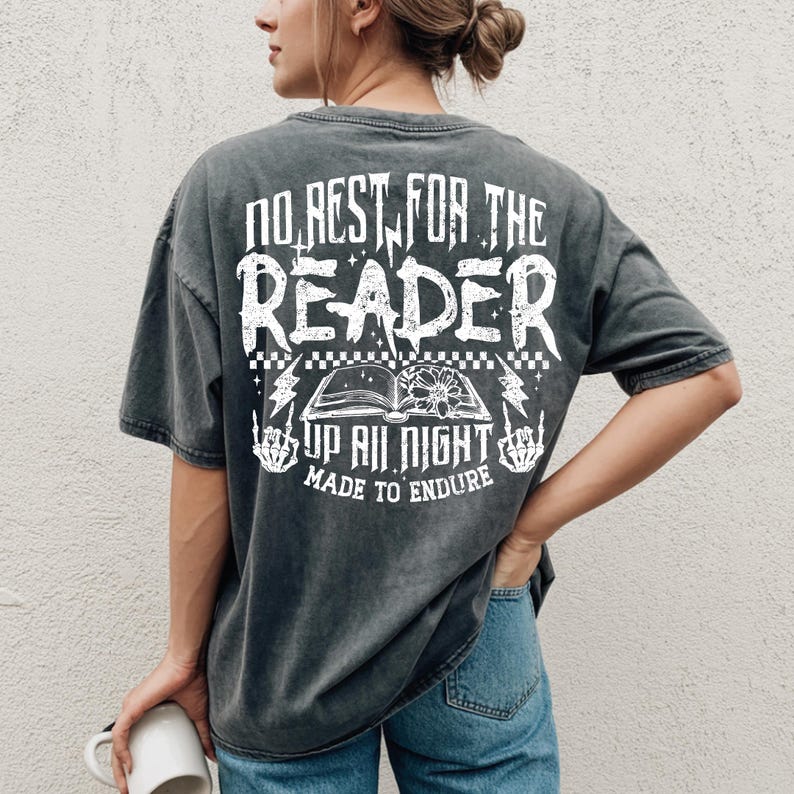 May include: Dark gray t-shirt with white text on the back. The text reads "NO REST FOR THE READER UP ALL NIGHT MADE TO ENDURE" with an open book graphic and lightning bolts. The shirt is worn by a person.