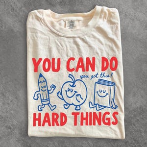May include: A cream-colored t-shirt featuring the red text YOU CAN DO HARD THINGS and a blue graphic of a smiling pencil, apple, and book with the blue text you got this! The shirt has a Comfort Colors brand label at the inner neckline.