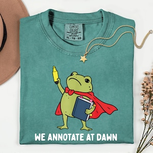 May include: A teal crewneck t-shirt featuring a graphic of a green frog wearing a red cape, holding a yellow highlighter and a blue book. The text "WE ANNOTATE AT DAWN" is printed in white below the frog. The shirt has a Comfort Colors brand label.