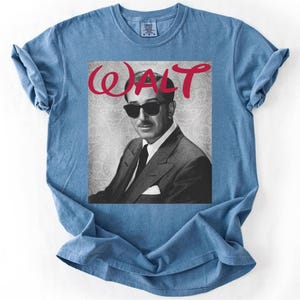 May include: A light blue t-shirt featuring a black and white image of a man wearing sunglasses and a suit. The word "WALT" is written in red cursive above the image. The shirt has a crew neck and short sleeves.