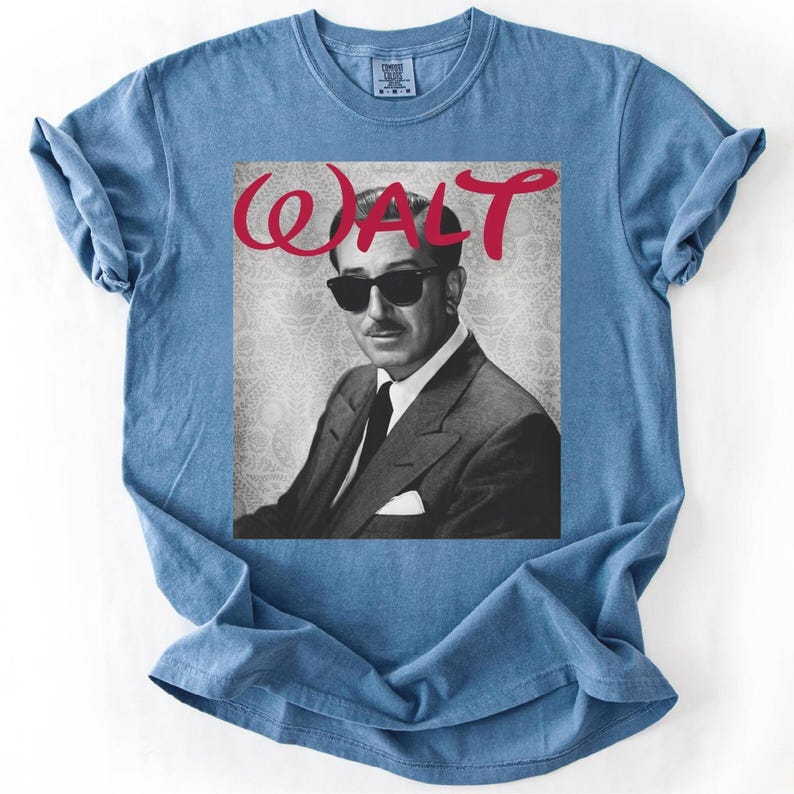 May include: A light blue t-shirt featuring a black and white image of a man wearing sunglasses and a suit. The word "WALT" is written in red cursive above the image. The shirt has a crew neck and short sleeves.