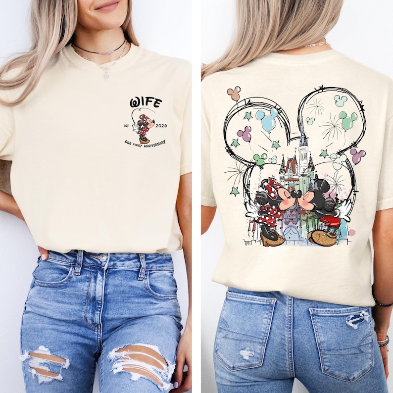 May include: Cream-colored t-shirt with a Mickey and Minnie Mouse design. The front features a small graphic with the word "WIFE" and "Our First Anniversary". The back has a larger design of the couple kissing in front of a castle, inside a Mickey Mouse head outline.
