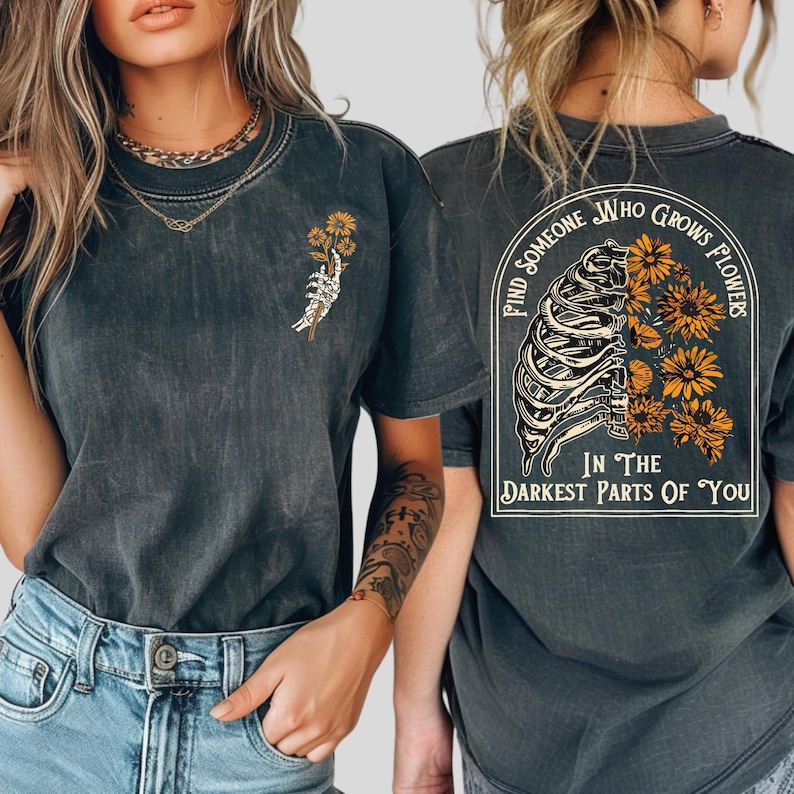 Puede incluir: Una camiseta gris carb&oacute;n con lavado &aacute;cido que presenta un peque&ntilde;o gr&aacute;fico de una mano esquel&eacute;tica sosteniendo flores amarillas en el frente, y un gr&aacute;fico grande en la espalda de una caja tor&aacute;cica con flores amarillas y el texto Find Someone Who Grows Flowers In The Darkest Parts Of You.