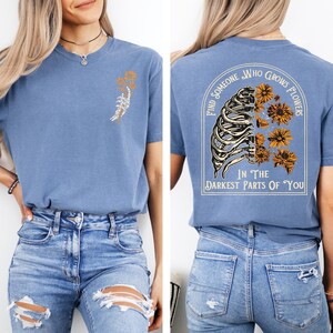 Puede incluir: Una camiseta azul con un peque&ntilde;o gr&aacute;fico en el pecho izquierdo de una mano de esqueleto sosteniendo flores amarillas. La parte posterior muestra un gr&aacute;fico grande de una caja tor&aacute;cica con flores amarillas y el texto, Find Someone Who Grows Flowers In The Darkest Parts Of You.