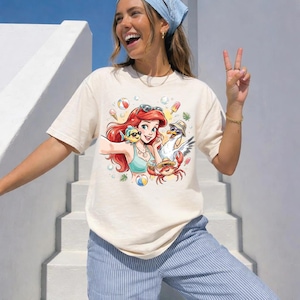May include: Cream-colored t-shirt featuring a cartoon illustration of a red-haired mermaid taking a selfie with a yellow fish, a seagull, and a crab. The design includes beach-themed elements like ice cream, beach balls, and sunglasses. The person in the image is wearing a blue bandana and blue striped pants.