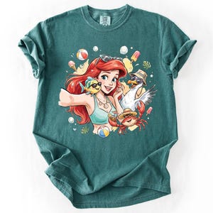 May include: Teal-colored t-shirt featuring a cartoon illustration of a red-haired woman with sunglasses, a fish, a seagull, and a crab, all wearing sunglasses and beach hats. The design includes beach balls, ice cream, and popsicles.