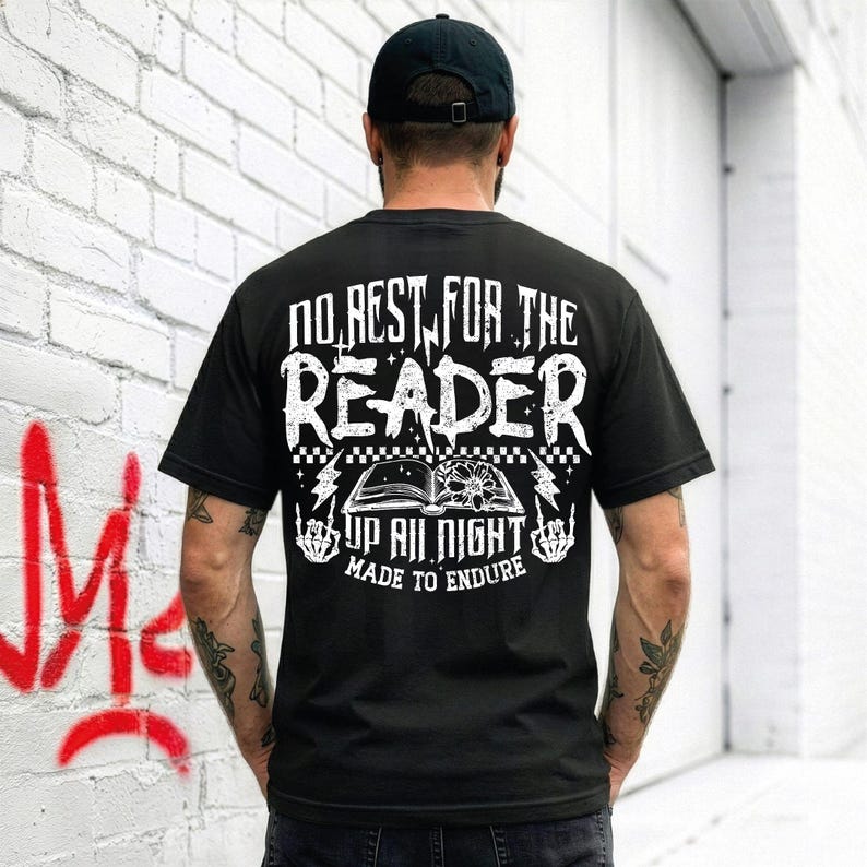 May include: Black t-shirt with a white graphic design on the back. The design includes the text "NO REST FOR THE READER" above an open book illustration, with the words "UP ALL NIGHT" and "MADE TO ENDURE" below. The person is wearing a black baseball cap.