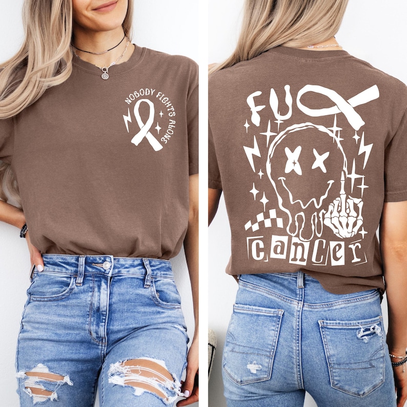 May include: A brown t-shirt featuring a white graphic on the front left chest of a ribbon with the text "NOBODY FIGHTS ALONE". The back displays a large white graphic of a melting smiley face, a skeleton hand, and the text "FUCK CANCER".