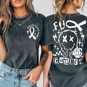 May include: A charcoal gray short-sleeve t-shirt featuring a white awareness ribbon and the text "NOBODY FIGHTS ALONE" on the front chest. The back displays a large white graphic of a melting smiley face, a skeleton hand, and "FUCK CANCER".