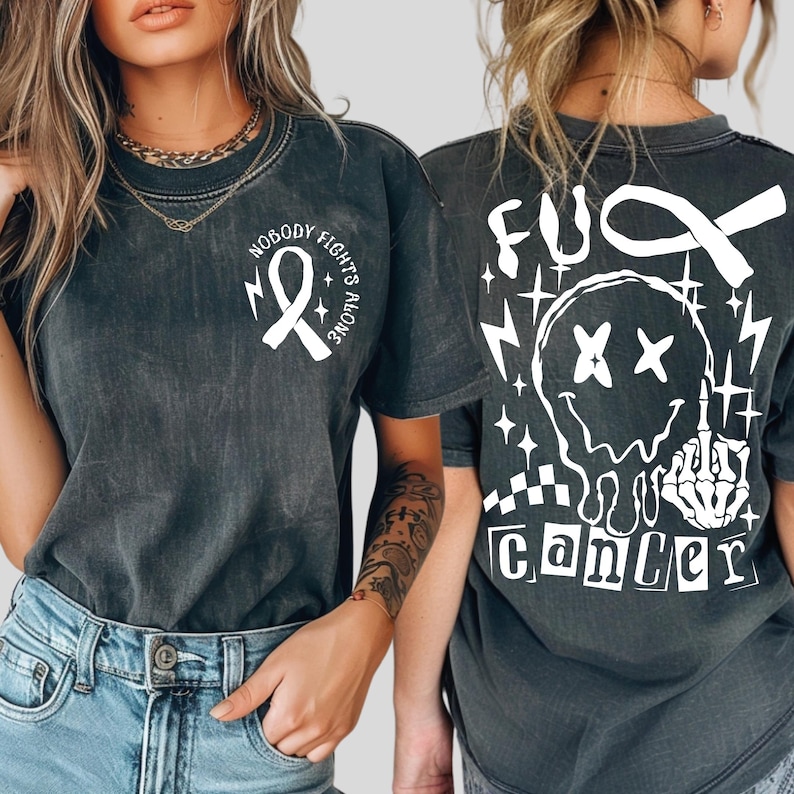 May include: A charcoal gray short-sleeve t-shirt featuring a white awareness ribbon and the text "NOBODY FIGHTS ALONE" on the front chest. The back displays a large white graphic of a melting smiley face, a skeleton hand, and "FUCK CANCER".