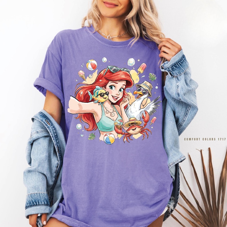 May include: A lilac-colored t-shirt featuring a colorful graphic of a cartoon character with red hair, sunglasses, and a turquoise bikini top. The graphic includes a fish, seagull, and crab, with beach-themed elements like ice cream and beach balls.