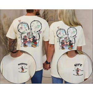 May include: Cream-coloured t-shirts with a Mickey and Minnie Mouse design kissing in front of a castle, with fireworks and a Mickey Mouse head outline. One shirt says "Husband" and the other says "Wife".