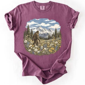 May include: A plum-colored short-sleeve t-shirt featuring a central graphic print of a brown Bigfoot creature walking through a field of colorful wildflowers, pine trees, and snow-capped mountains under a blue sky with white clouds.