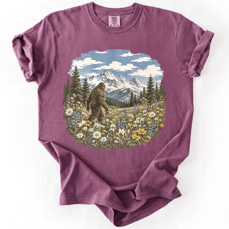 May include: A plum-colored short-sleeve t-shirt featuring a central graphic print of a brown Bigfoot creature walking through a field of colorful wildflowers, pine trees, and snow-capped mountains under a blue sky with white clouds.