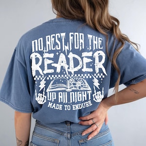 May include: Blue t-shirt with white graphic text: "NO REST FOR THE READER" above an open book illustration. Below the book are the words "UP ALL NIGHT MADE TO ENDURE". Lightning bolts and skeleton hands flank the book.
