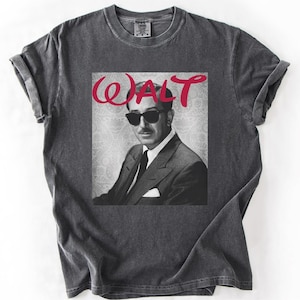 May include: Dark gray t-shirt featuring a black and white image of a man wearing sunglasses, with the red word "WALT" above the image. The shirt has rolled-up sleeves and a crew neck.