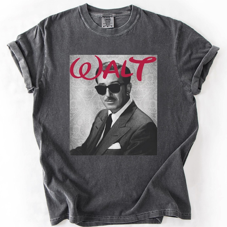 May include: Dark gray t-shirt featuring a black and white image of a man wearing sunglasses, with the red word "WALT" above the image. The shirt has rolled-up sleeves and a crew neck.