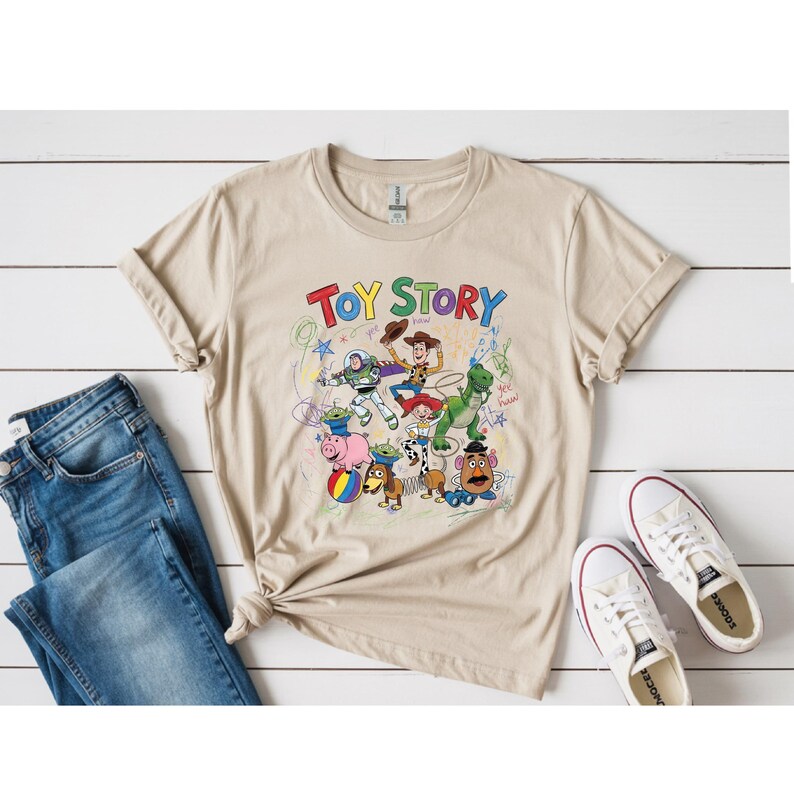 May include: Beige t-shirt featuring the words "Toy Story" above a colorful graphic of characters from the animated film. The shirt is paired with blue jeans and white sneakers with red accents, set against a white wood background.