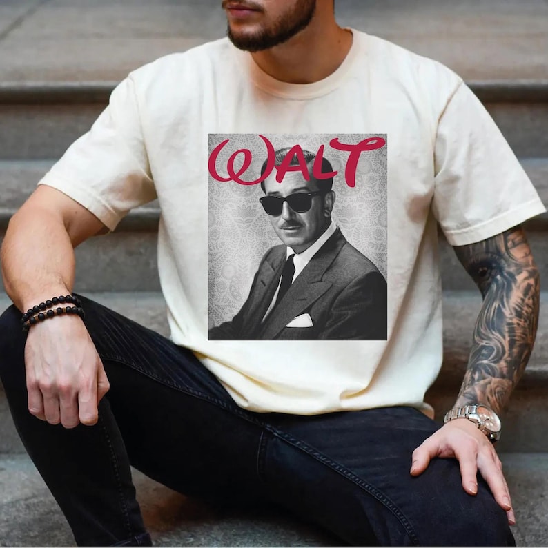 May include: Cream-colored t-shirt featuring a black and white image of a man wearing sunglasses and a suit, with the name "WALT" in red script above the image. The shirt is a casual style.