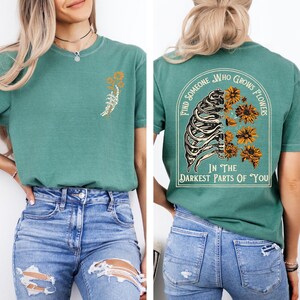 Puede incluir: Una camiseta de manga corta color verde azulado con un peque&ntilde;o gr&aacute;fico de una mano esquel&eacute;tica sosteniendo flores en el pecho izquierdo y un gr&aacute;fico grande en la espalda de una caja tor&aacute;cica con flores y el texto Find Someone Who Grows Flowers In The Darkest Parts Of You.