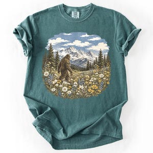 May include: A teal short-sleeve t-shirt featuring a circular graphic print of a brown Bigfoot creature walking through a field of wildflowers, with tall pine trees and snow-capped mountains in the background under a blue sky with white clouds.