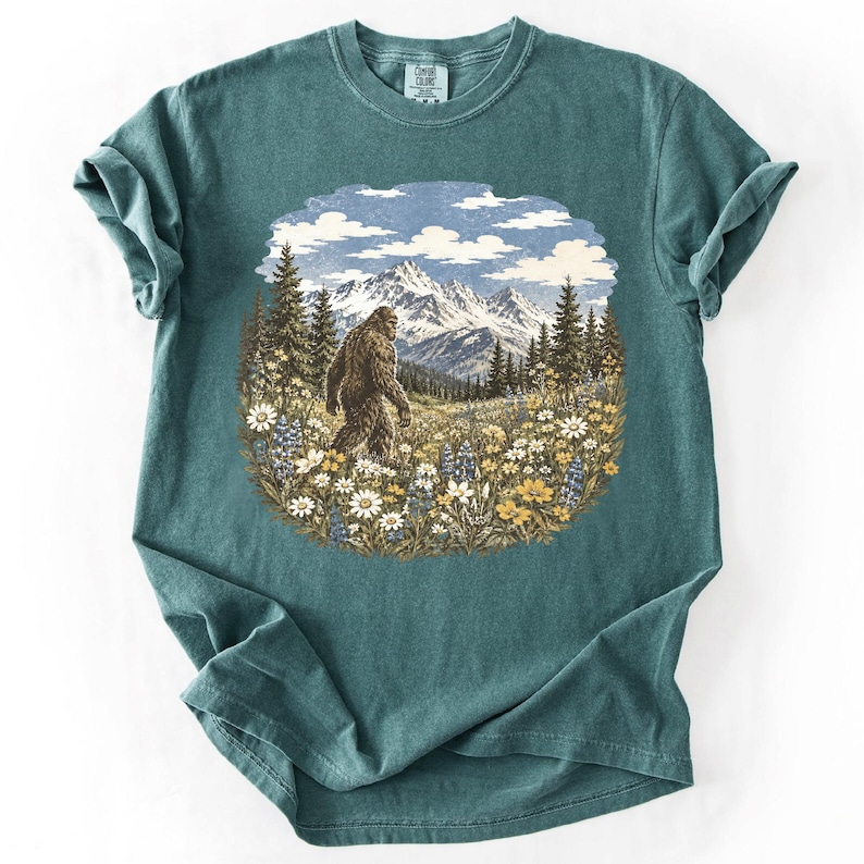 May include: A teal short-sleeve t-shirt featuring a circular graphic print of a brown Bigfoot creature walking through a field of wildflowers, with tall pine trees and snow-capped mountains in the background under a blue sky with white clouds.