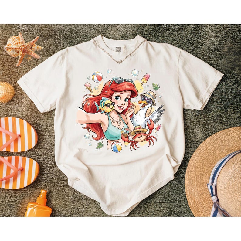 May include: Off-white t-shirt featuring a cartoon illustration of a red-haired woman taking a selfie with a fish, seagull, and crab. The design includes beach balls, ice cream, and sunglasses. A straw hat, flip-flops, and sunscreen are also visible.