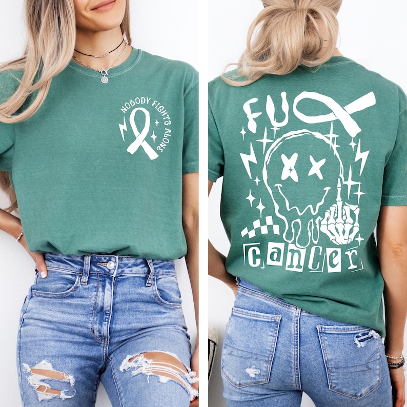 May include: A green short-sleeve t-shirt featuring a white graphic on the front chest of a ribbon with the text "NOBODY FIGHTS ALONE." The back displays a large white graphic of a melting smiley face, a ribbon, and the text "FUCK CANCER."