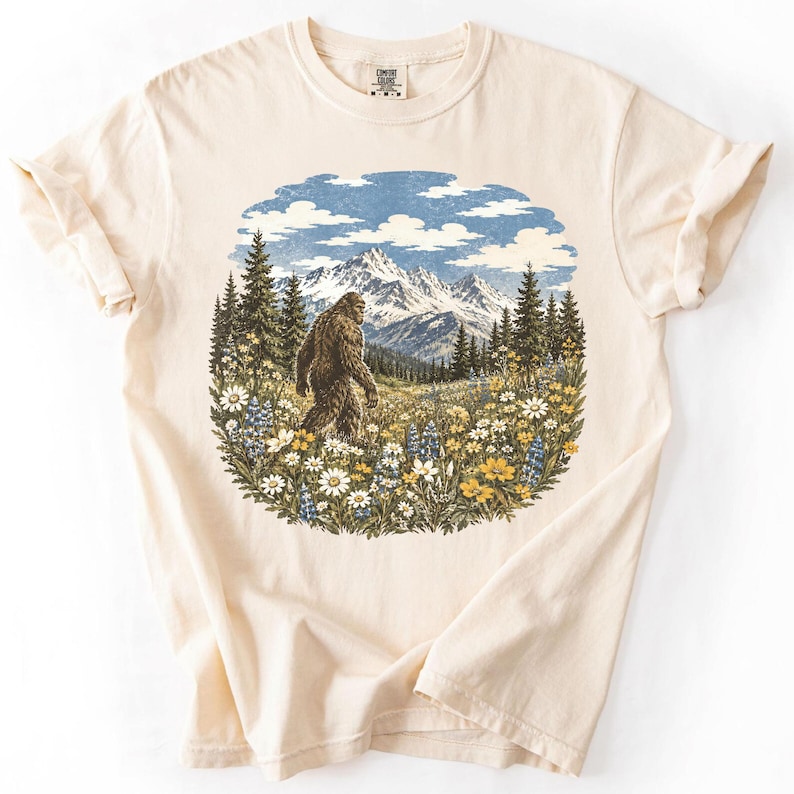 May include: A cream-colored short-sleeve t-shirt featuring a circular graphic print of a brown Bigfoot walking through a field of white and yellow wildflowers, with a backdrop of green pine trees and snow-capped mountains under a blue sky.