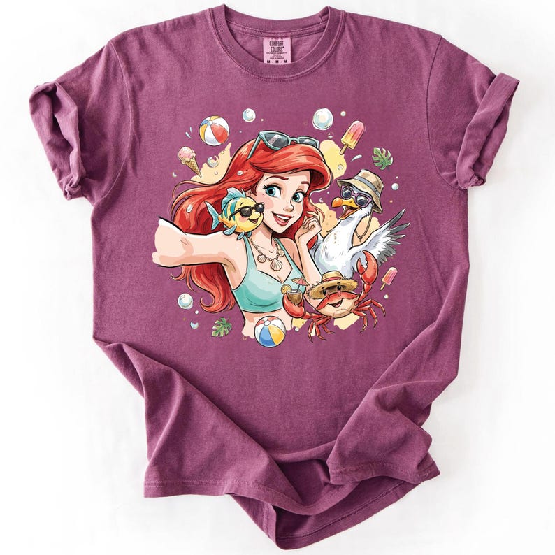 May include: A maroon t-shirt featuring a colorful graphic of a cartoon character with red hair, a blue top, and sunglasses, posing with a fish, crab, and seagull. The design includes beach balls, ice cream, and bubbles.