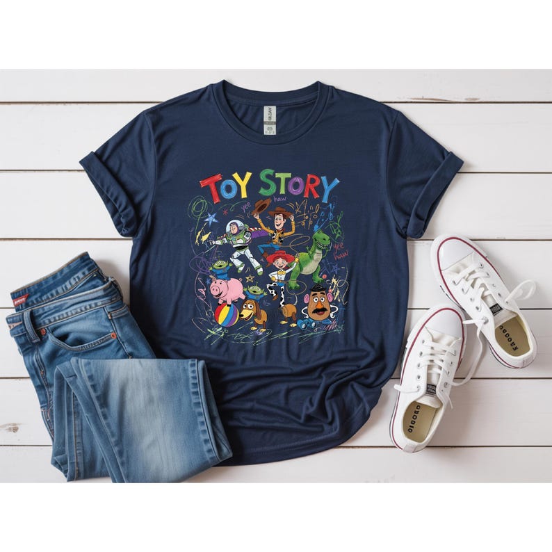 May include: Navy blue t-shirt featuring the words "TOY STORY" in colorful letters, with a graphic of characters from the movie. The shirt is paired with blue jeans and white sneakers, all arranged on a white wooden surface.