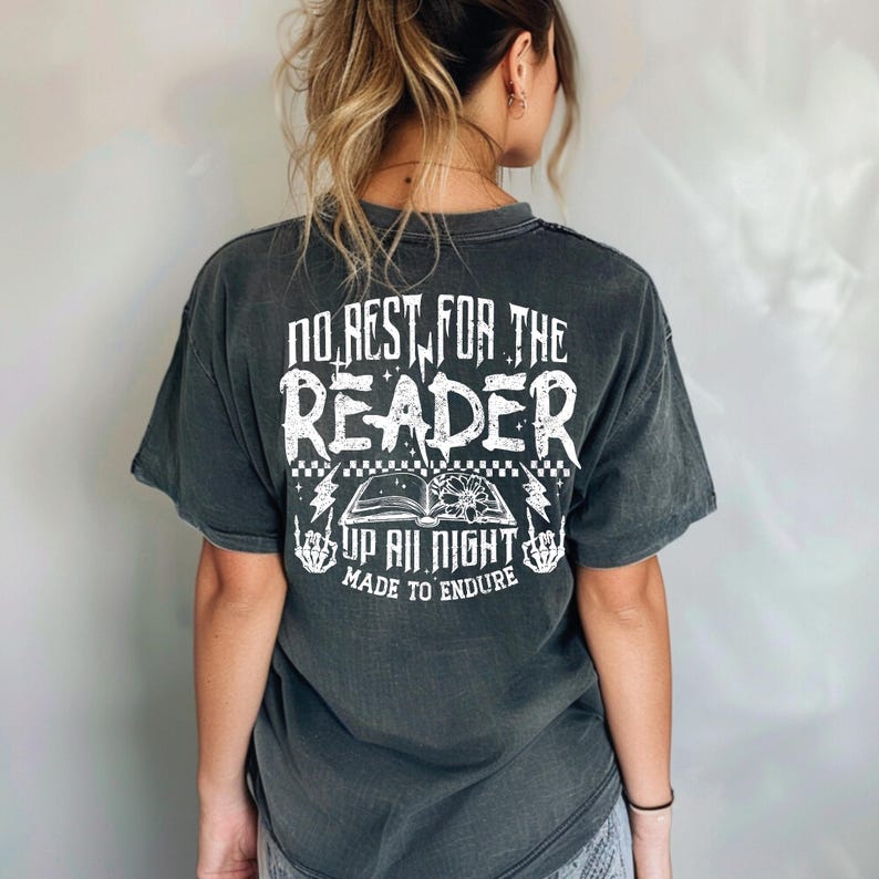 May include: Dark grey t-shirt with white text and graphics. The text reads "NO REST FOR THE READER" above an open book graphic with lightning bolts and the words "UP ALL NIGHT MADE TO ENDURE".