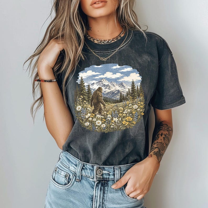 May include: A charcoal gray short-sleeve t-shirt featuring a circular graphic print of a Bigfoot creature standing in a field of wildflowers with a mountain range and pine trees in the background under a blue sky with white clouds.