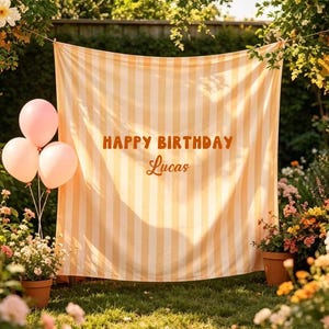 May include: A striped orange and white backdrop with the words "HAPPY BIRTHDAY Lucas" is strung between trees. Three pink balloons are tied together on the left. The scene is set in a garden with flowers and potted plants.