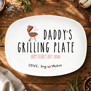 May include: An oval white ceramic serving plate featuring a red charcoal grill graphic and black text that reads DADDY'S GRILLING PLATE, HAPPY FATHER'S DAY! 2026, and LOVE, IVY [heart symbol] MILES printed in the center of the plate surface.