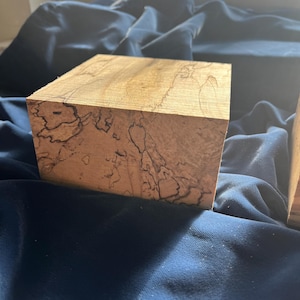 May include: A close-up of a wooden cube with a light brown color and dark, intricate patterns. The cube is sitting on a dark blue fabric. The wood grain is visible, and the cube has a smooth, polished surface.