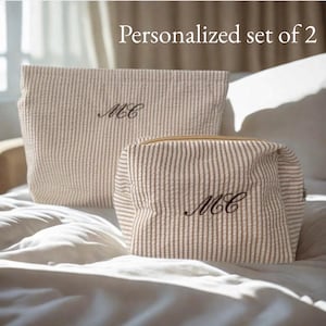 May include: Two beige and white striped cosmetic bags with the text "Personalized set of 2". Each bag is embroidered with the initials "MC" in black. One bag is rectangular, and the other is a smaller, zippered pouch.