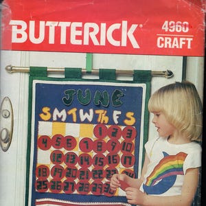 May include: A vintage Butterick craft pattern for a felt calendar. The calendar is blue with red and yellow accents, featuring the month of June and a grid for the days. The word "Jennifer" is on the bottom. A child is standing next to the calendar.