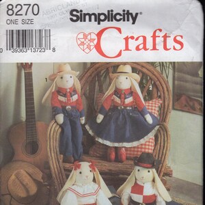 Puede incluir: Patrón de Simplicity Crafts con cuatro muñecos de conejos vestidos con trajes de vaquero y vaquera. Los muñecos llevan sombreros, camisas, faldas y pantalones en rojo, blanco y azul. El patrón incluye instrucciones.