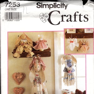 Puede incluir: Un patrón de costura de Simplicity Crafts para hacer cinco accesorios de cocina diferentes con temática de conejito. El patrón incluye instrucciones para hacer un porta-toallas de cocina con forma de conejito, un guante de horno con forma de conejito, una decoración de pared con forma de conejito, un tope de puerta con forma de conejito y una toalla de cocina con forma de conejito. El patrón está impreso en un fondo blanco con un borde rosa.