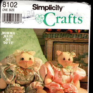 May include: Simplicity Crafts pattern featuring two plush bear dolls. The bears have peach-colored faces, ears, and feet, with blush-colored cheeks. They wear pastel-colored outfits with decorative trim. The text "MOMMA MADE ME DO IT!" is visible.