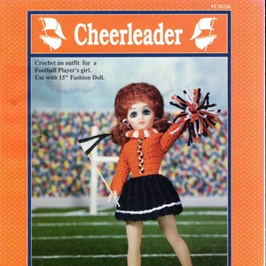 May include: A doll wearing a crocheted cheerleader outfit, including an orange sweater, navy blue skirt, and white shoes. The doll is holding a pom-pom and a flag. The image is from a Fibre-Craft pattern for a 15" fashion doll.