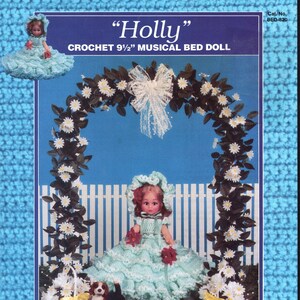 Könnte beinhalten: Ein Häkelmuster-Cover mit einer Puppe in einem hellblauen Kleid, einem kleinen Hund und Blumendekorationen. Der Text lautet "Holly" und "CROCHET 24 cm MUSICAL BED DOLL". Der Hintergrund ist ein blau-weißes Design.