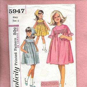 May include: Simplicity sewing pattern number 5947 for a child's dress. The pattern includes three different styles of dresses, one with a gingham print, one with a floral print, and one with a solid pink color. The pattern also includes instructions for making a smocked trim.