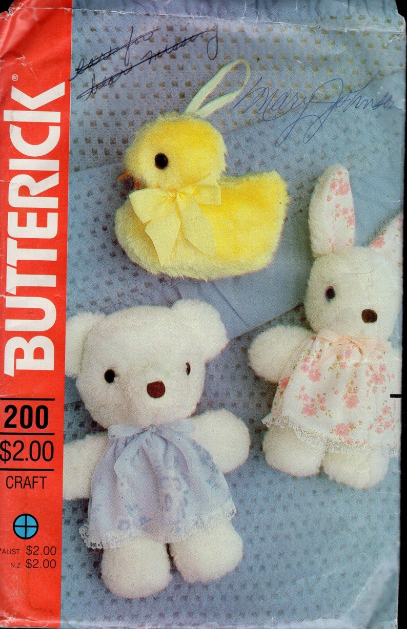 May include: A Butterick 200 craft pattern envelope featuring a yellow plush chick with a ribbon, a white plush bear wearing a light blue dress with lace trim, and a white plush rabbit wearing a floral print dress with lace trim.