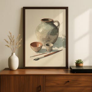 May include: A framed wall art print featuring a still life illustration of a large, light green ceramic pitcher, a small terracotta bowl, a gray cup, and two paintbrushes arranged on a light-colored surface with soft shadows.