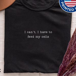 May include: A black t-shirt featuring the white embroidered text I can't, I have to feed my cells. The shirt includes a tag at the neckline and a circular blue, white, and red badge in the upper right corner that reads Designed & Printed in USA.