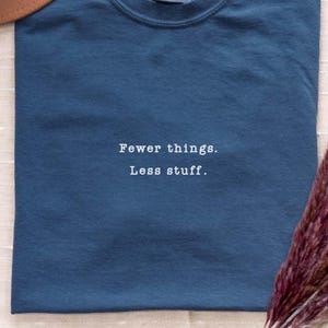 Könnte beinhalten: Ein dunkelblaues Kurzarm-T-Shirt mit dem in Weiß gestickten Text "Fewer things. Less stuff." mittig auf der Brust. Das Shirt hat einen Rundhalsausschnitt und ein sichtbares Markenetikett an der Innenseite des Kragens.