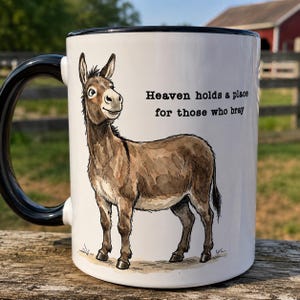 May include: A white ceramic mug with a black handle and rim, featuring a watercolor-style illustration of a brown donkey standing on a light brown patch of ground. The text "Heaven holds a place for those who bray" is printed in black.