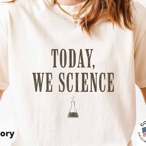 May include: An ivory short-sleeved t-shirt featuring the dark brown text "TODAY, WE SCIENCE" printed on the front. Below the text is a small graphic of a laboratory flask with bubbles rising from the top. The shirt has a crew neckline.